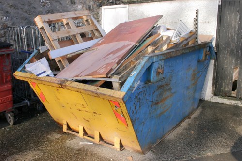 A large, open-top industrial skips-style container with a yellow front panel and blue sides, filled with various discarded wooden pallets, particle boards, and construction debris. The wooden pallets are weathered and partly broken, with some lying flat and others leaning against each other. The particle boards are of different sizes, with some showing chipped edges and rough textures. The container is situated on an outdoor paved surface beside a grey wall and a weathered wooden gate, with a black fence partially visible in the background.Natural sunlight illuminates the scene, casting soft shadows around the container. This setting is reminiscent of a waste collection or rubbish removal area that could be typical for businesses in Barnet, where Business Waste Removal Barnet offers rubbish disposal services. The surrounding environment suggests a typical industrial or commercial loading or waste storage zone, emphasizing the types of bulk materials handled in rubbish removal operations, aligned with the company's service focus in the local area.