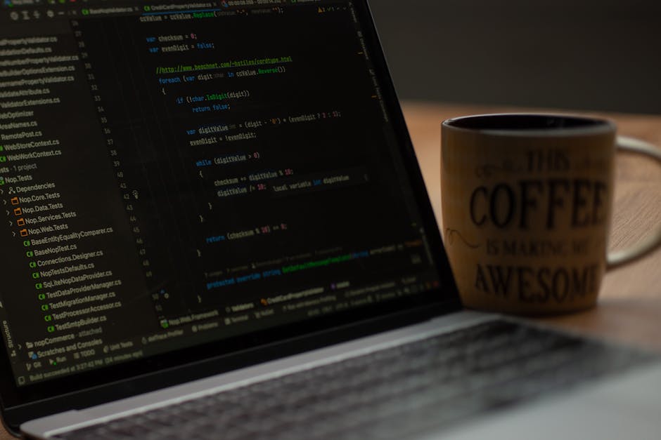 A person working on a MacBook laptop with a dark screen displaying lines of code and text in an integrated development environment. The laptop is positioned on a dark reflective surface, with natural 