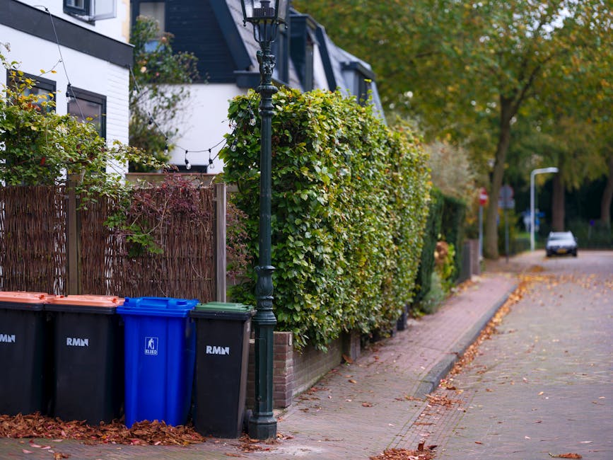 A person wearing a white glove and a wristwatch is holding open a large white woven polypropylene sack filled with garden waste, including small leafy branches with green leaves and some dried brown f